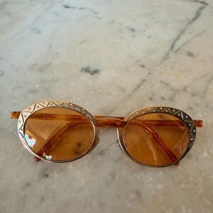 Op de afbeelding: Ovale zonnebril met amberkleurige glazen en goudkleurige metalen monturen. De randen zijn versierd met een gegraveerd geometrisch patroon en kleine ingebedde kristallen. Het binnenframe is gemarkeerd met de tekst 24 KT GOLD PLATED.