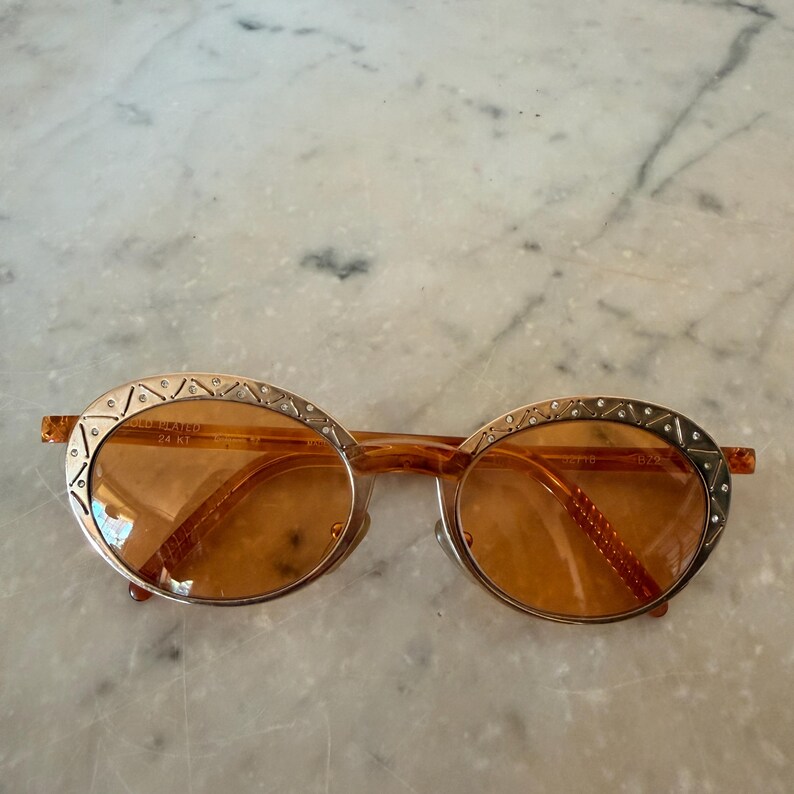 Op de afbeelding: Ovale zonnebril met amberkleurige glazen en goudkleurige metalen monturen. De randen zijn versierd met een gegraveerd geometrisch patroon en kleine ingebedde kristallen. Het binnenframe is gemarkeerd met de tekst 24 KT GOLD PLATED.