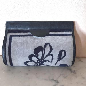 Può includere: Una pochette rettangolare con un motivo floreale. La borsa presenta un tessuto grigio con un motivo floreale e finiture blu scuro. La parte superiore della pochette è in pelle blu scuro. La borsa è su una superficie bianca.
