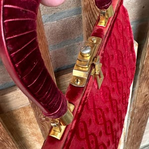 Pu&ograve; includere: Una borsa in velluto rosso intenso con un manico strutturato e arrotondato, rivestito in velluto rosso abbinato. La borsa presenta una struttura in metallo dorato con una chiusura sicura e un motivo in rilievo ripetuto sulla superficie.