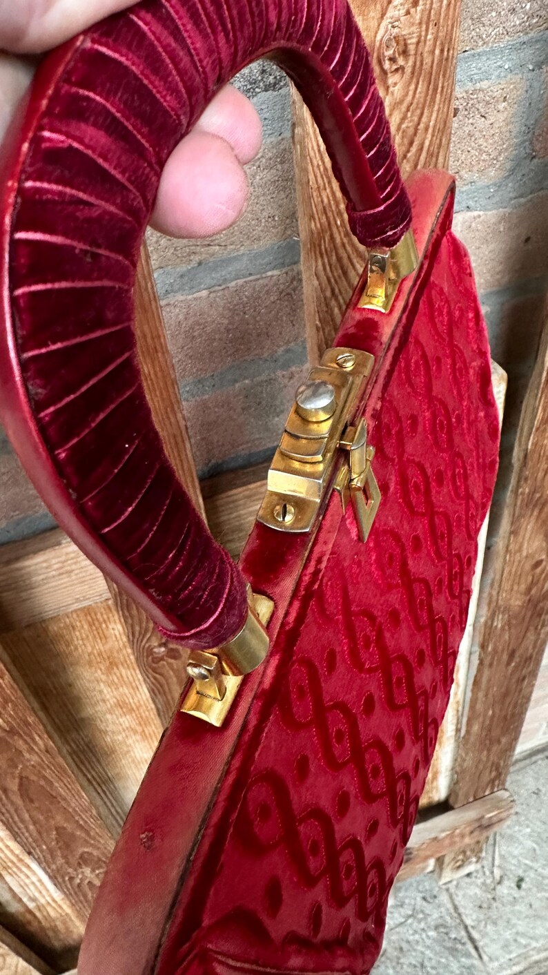 Pu&ograve; includere: Una borsa in velluto rosso intenso con un manico strutturato e arrotondato, rivestito in velluto rosso abbinato. La borsa presenta una struttura in metallo dorato con una chiusura sicura e un motivo in rilievo ripetuto sulla superficie.