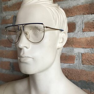 May include: Gold-tone eyeglasses with a blue top bar and temples. The lenses are clear, and the brand name "Silhouette" is visible on the lens. The glasses are displayed on a white mannequin head against a brick wall.