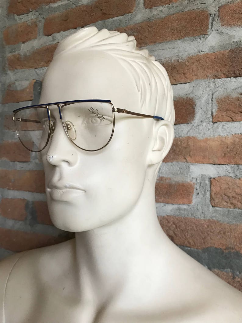May include: Gold-tone eyeglasses with a blue top bar and temples. The lenses are clear, and the brand name "Silhouette" is visible on the lens. The glasses are displayed on a white mannequin head against a brick wall.
