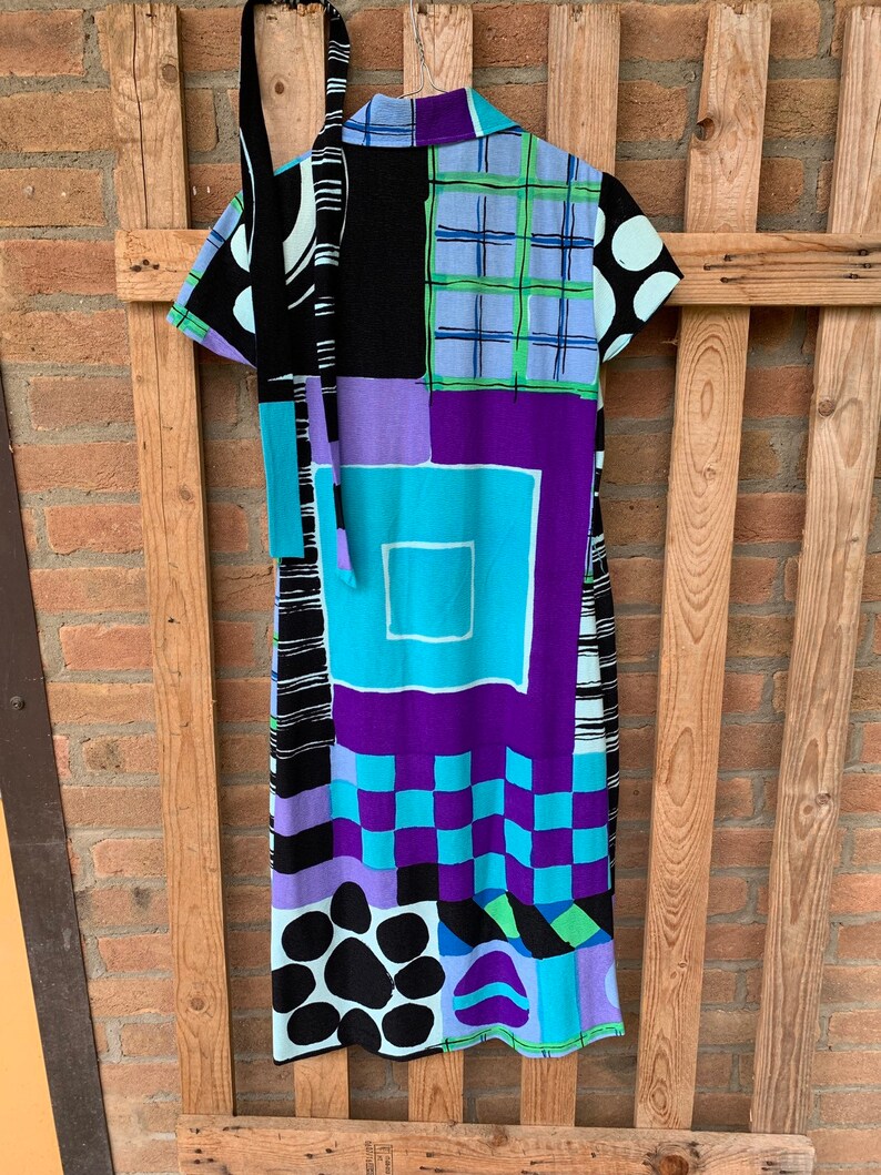 Puede incluir: Un vestido estampado con un dise&ntilde;o geom&eacute;trico colorido en negro, blanco, azul, verde y morado. El vestido tiene manga corta y un lazo en el cuello.