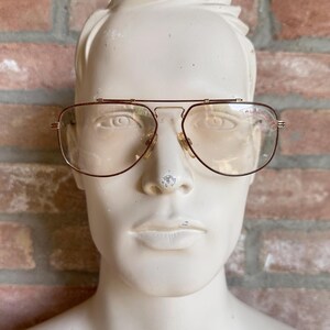 Può includere: Occhiali in stile aviatore con una sottile montatura in metallo color oro e un design a doppio ponte. Le lenti sono chiare e hanno una tinta sottile. La parola "ALFA" è stampata in rosso sulla lente superiore destra. Gli occhiali sono esposti su un manichino.