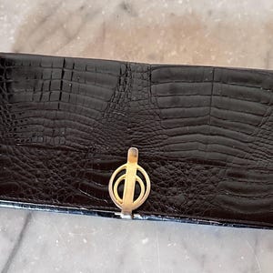 Könnte beinhalten: Eine schwarze, rechteckige Geldbörse mit Krokodilhautstruktur. Die Geldbörse hat einen goldfarbenen Verschluss in der Mitte. Die Geldbörse ist geschlossen und scheint aus Leder zu sein.