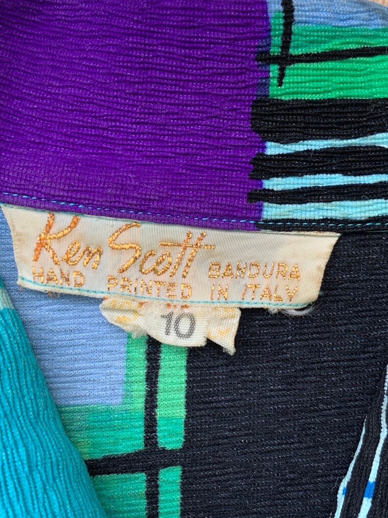 Puede incluir: Un primer plano de una etiqueta de ropa con el texto "Ken Scott Bandura Hand Printed in Italy" y el n&uacute;mero "10". La etiqueta est&aacute; unida a una tela con estampado azul, verde, morado y negro.