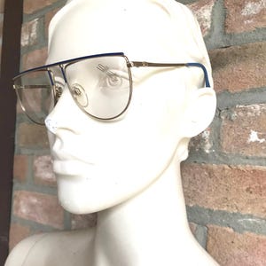 May include: Gold-rimmed eyeglasses with a blue top bar and temple tips. The lenses are clear, and the brand name "Silhouette" is visible on the lens. The glasses are displayed on a white mannequin head.