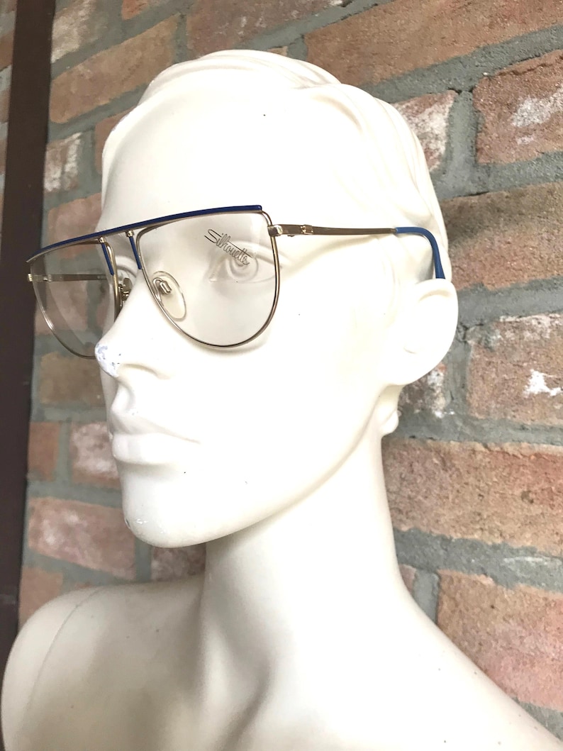 May include: Gold-rimmed eyeglasses with a blue top bar and temple tips. The lenses are clear, and the brand name "Silhouette" is visible on the lens. The glasses are displayed on a white mannequin head.