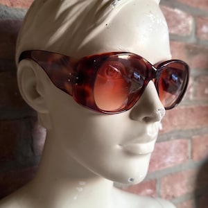 May include: Oval-shaped sunglasses with a tortoiseshell pattern and gradient lenses. The frames have a dark brown and amber color scheme. The sunglasses are displayed on a mannequin head against a brick wall background.