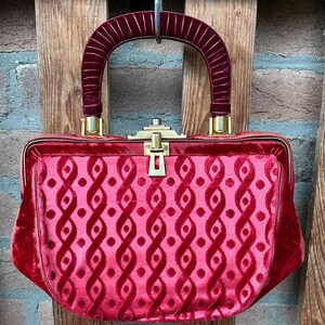 Pu&ograve; includere: Una borsa a mano in velluto rosso intenso con una struttura rigida e una chiusura in metallo dorato. L'esterno presenta un motivo ripetitivo di nastri verticali intrecciati e piccoli punti. &Egrave; dotata di un manico arcuato in velluto a coste.