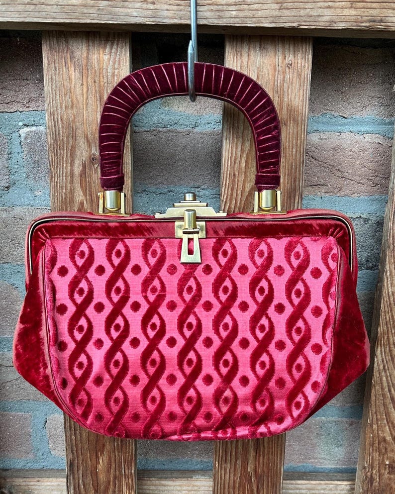 Pu&ograve; includere: Una borsa a mano in velluto rosso intenso con una struttura rigida e una chiusura in metallo dorato. L'esterno presenta un motivo ripetitivo di nastri verticali intrecciati e piccoli punti. &Egrave; dotata di un manico arcuato in velluto a coste.