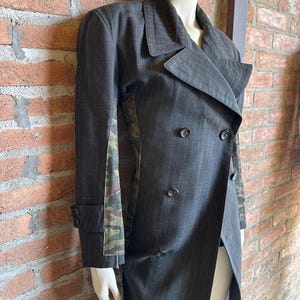 May include: A dark gray pinstripe double-breasted coat with a unique design. The coat features a camouflage pattern on the side panels and a long, asymmetrical hemline. The coat is displayed on a mannequin.