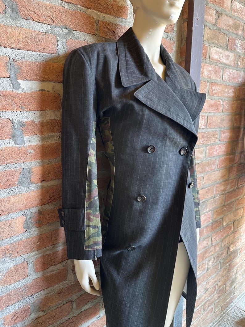 May include: A dark gray pinstripe double-breasted coat with a unique design. The coat features a camouflage pattern on the side panels and a long, asymmetrical hemline. The coat is displayed on a mannequin.