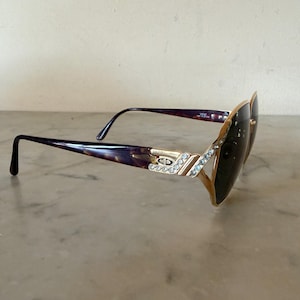 May include: Tortoiseshell sunglasses with dark lenses and gold-tone metal accents. The temples feature a gold-tone logo plate with the letters CD and a row of clear crystals. The frame front also includes a decorative row of clear crystals.