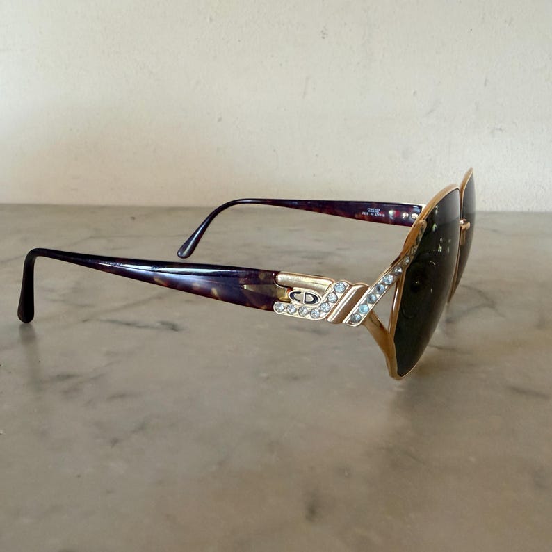 May include: Tortoiseshell sunglasses with dark lenses and gold-tone metal accents. The temples feature a gold-tone logo plate with the letters CD and a row of clear crystals. The frame front also includes a decorative row of clear crystals.