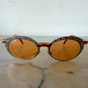 Op de afbeelding: Ovaalvormige zonnebril met oranje getinte glazen en een metalen montuur. Het montuur is versierd met een herhalend driehoekspatroon en kleine ingebedde heldere kristallen langs de buitenrand, verbonden door een neusbrug en dunne pootjes.