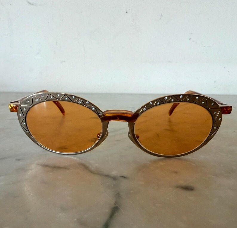 Op de afbeelding: Ovaalvormige zonnebril met oranje getinte glazen en een metalen montuur. Het montuur is versierd met een herhalend driehoekspatroon en kleine ingebedde heldere kristallen langs de buitenrand, verbonden door een neusbrug en dunne pootjes.