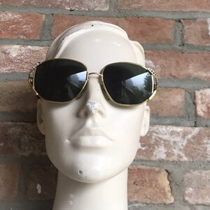 May include: Gold-tone sunglasses featuring dark, tinted lenses and a thin metal frame. The temples are accented with small, clear decorative stones embedded along the sides near the hinges, providing a subtle sparkle to the eyewear design.
