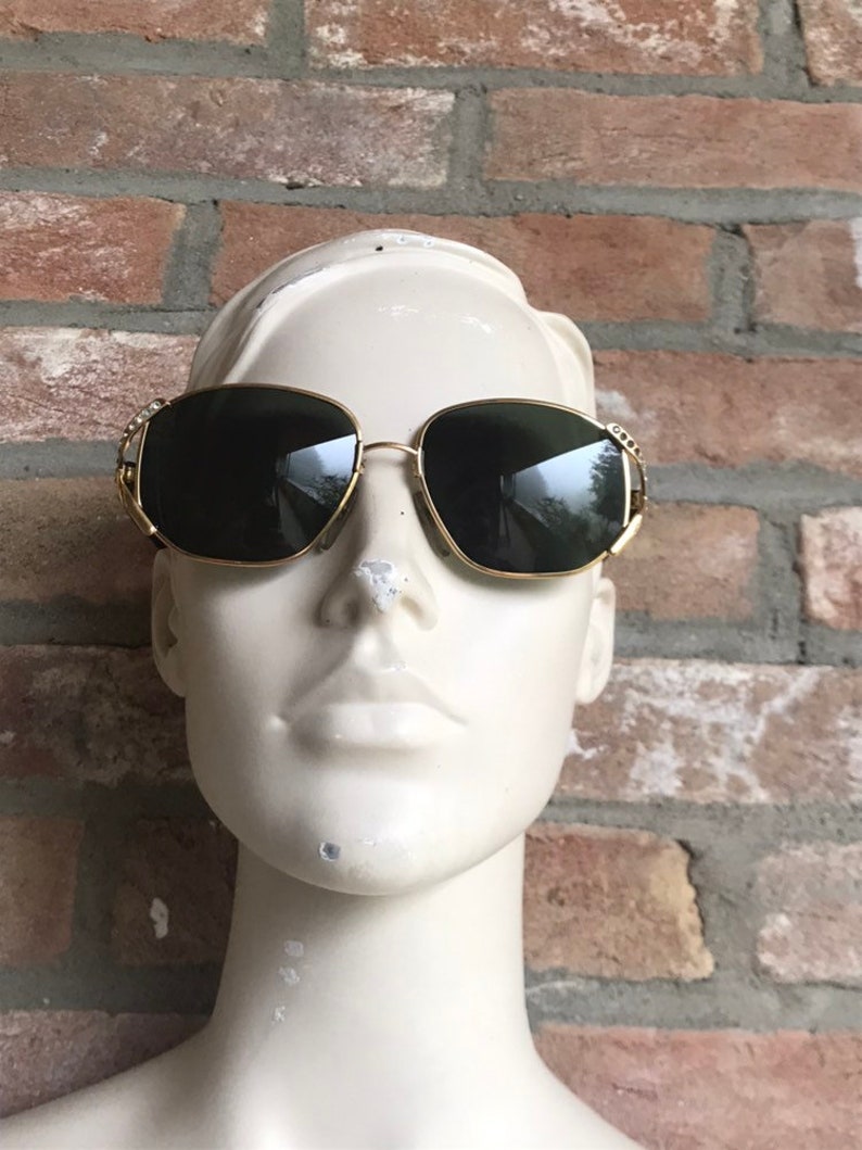 May include: Gold-tone sunglasses featuring dark, tinted lenses and a thin metal frame. The temples are accented with small, clear decorative stones embedded along the sides near the hinges, providing a subtle sparkle to the eyewear design.