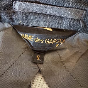 May include: Close-up of a dark gray blazer interior with a black label. The label reads "COMME des GARCONS" in gold lettering, with a small "S" size tag. The blazer's fabric has a subtle plaid pattern.