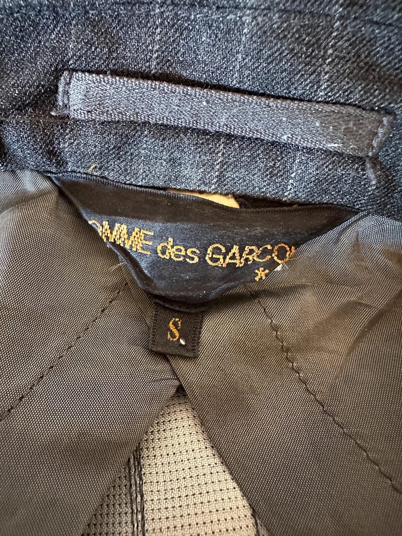 May include: Close-up of a dark gray blazer interior with a black label. The label reads "COMME des GARCONS" in gold lettering, with a small "S" size tag. The blazer's fabric has a subtle plaid pattern.