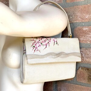 Può includere: Una borsa color crema con un ramo di ciliegio dipinto a mano con fiori rosa e steli marroni sulla patta anteriore. La borsa include un singolo manico superiore e una fascia decorativa in tessuto orizzontale al centro della patta.