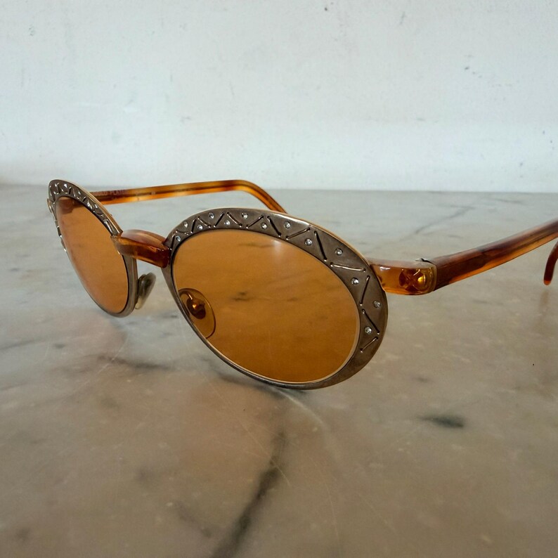 Op de afbeelding: Ovale zonnebril met amberkleurige glazen en een bruin montuur met schildpadpatroon. De metalen randen rond de glazen hebben een matte goudkleurige afwerking en zijn versierd met een geometrisch patroon en kleine ingebedde kristallen.