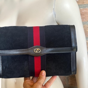 Può includere: Pochette in camoscio nero con una striscia rossa e verde al centro. La pochette ha un cinturino in pelle con un'emblema color oro. I bordi sono rifiniti in pelle nera.
