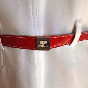 Puede incluir: Primer plano de una prenda blanca con un cintur&oacute;n rojo brillante. El cintur&oacute;n tiene un acabado brillante y costuras blancas. Una hebilla cuadrada plateada con un logotipo est&aacute; centrada en el cintur&oacute;n. La prenda tiene costuras verticales.