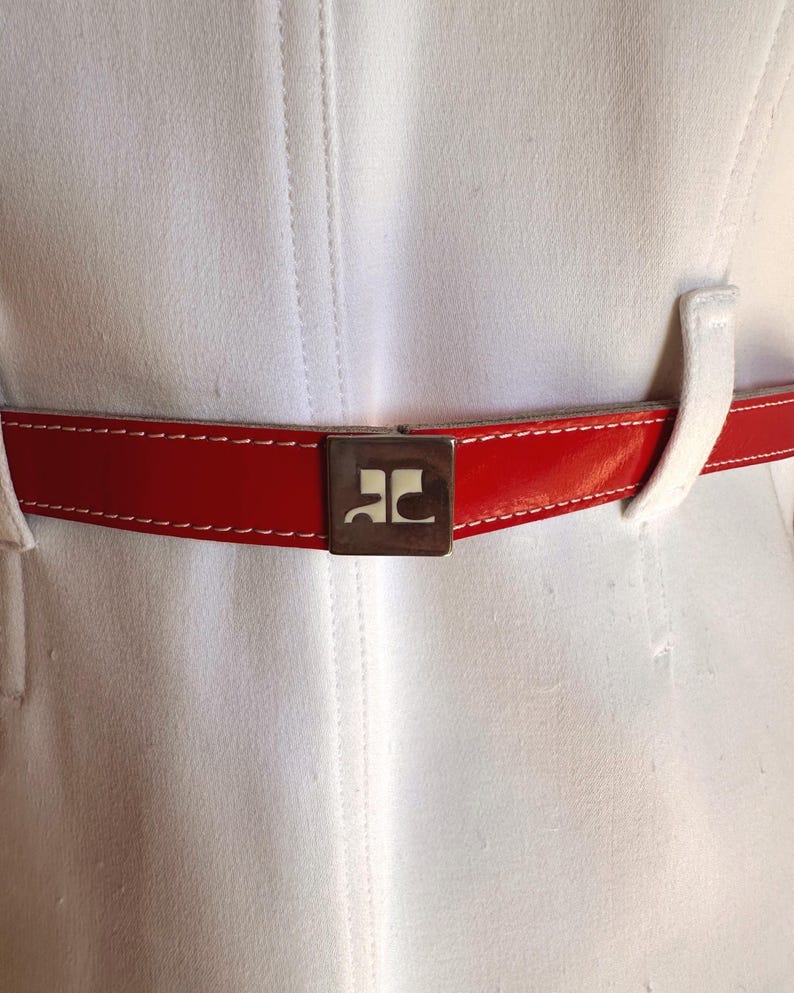 Puede incluir: Primer plano de una prenda blanca con un cintur&oacute;n rojo brillante. El cintur&oacute;n tiene un acabado brillante y costuras blancas. Una hebilla cuadrada plateada con un logotipo est&aacute; centrada en el cintur&oacute;n. La prenda tiene costuras verticales.