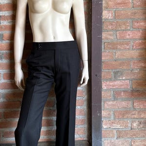 Peut inclure: Pantalon noir &agrave; fines rayures et jambes larges, pr&eacute;sent&eacute; sur un mannequin. Le pantalon taille haute est ferm&eacute; par un bouton. Le mannequin est devant un mur de briques.