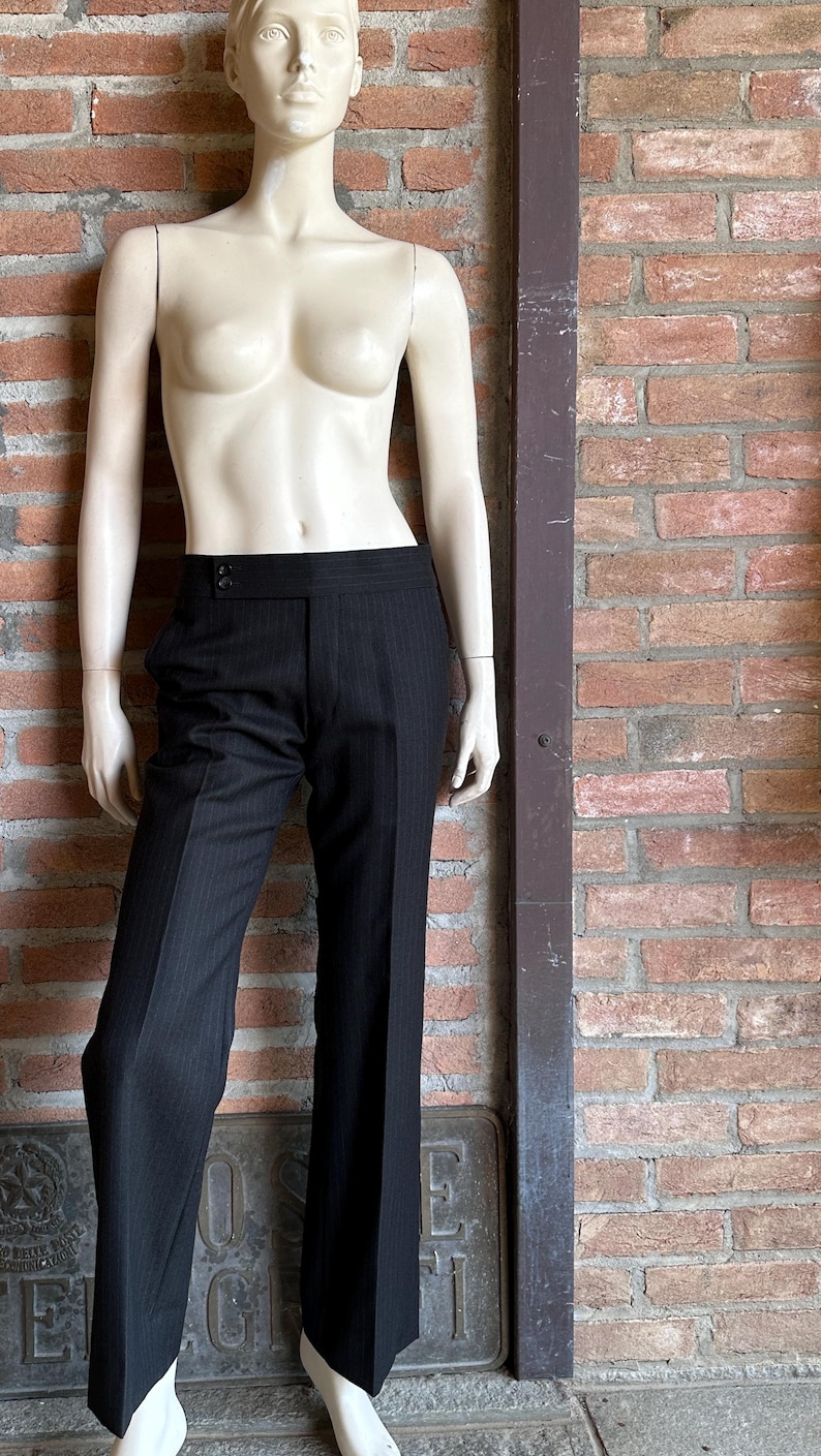 Peut inclure: Pantalon noir &agrave; fines rayures et jambes larges, pr&eacute;sent&eacute; sur un mannequin. Le pantalon taille haute est ferm&eacute; par un bouton. Le mannequin est devant un mur de briques.