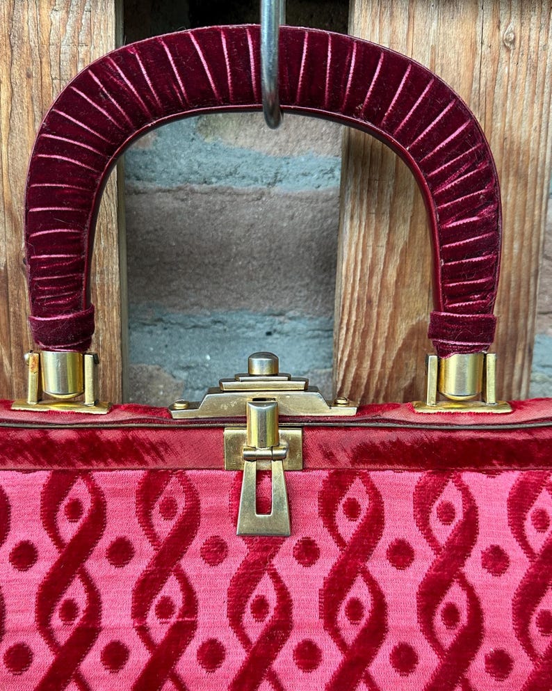 Pu&ograve; includere: Una borsa con manico in velluto rosso intenso con nervature verticali e corpo in velluto abbinato con un motivo ripetitivo di anelli e punti in velluto in rilievo. La borsa include una chiusura e finiture in metallo color oro.