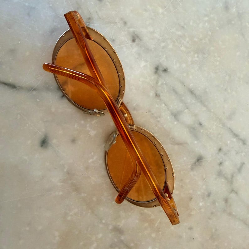 Op de afbeelding: Ronde zonnebril met amberkleurige glazen en een transparant oranje montuur. De ronde glazen zijn gevat in een dunne metalen rand met kleine decoratieve studs langs de buitenrand. De pootjes zijn gemaakt van bijpassend oranje materiaal.