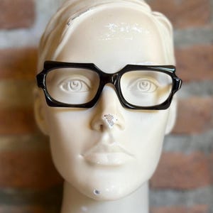 Könnte beinhalten: Schwarze Brille mit einem dicken, kantigen Rahmendesign und spitz zulaufenden oberen Ecken. Die Brille befindet sich auf einem glatten, hellen Schaufensterpuppenkopf und zeigt die markante, geometrische Form der Ränder sowie die klaren Gläser.