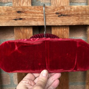 Pu&ograve; includere: Una borsa in velluto rosso intenso caratterizzata da una consistenza morbida e vellutata e da una forma ovale allungata e strutturata. La borsa &egrave; appesa a una gruccia di metallo, mettendo in risalto il suo colore cremisi ricco e saturo.