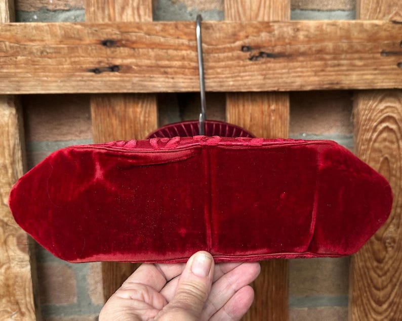Pu&ograve; includere: Una borsa in velluto rosso intenso caratterizzata da una consistenza morbida e vellutata e da una forma ovale allungata e strutturata. La borsa &egrave; appesa a una gruccia di metallo, mettendo in risalto il suo colore cremisi ricco e saturo.