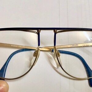 May include: Gold-rimmed eyeglasses with a blue top bar and blue temples. The lenses are clear, and the frame has a distinctive aviator shape. The brand name "Silhouette" is visible on the lens.