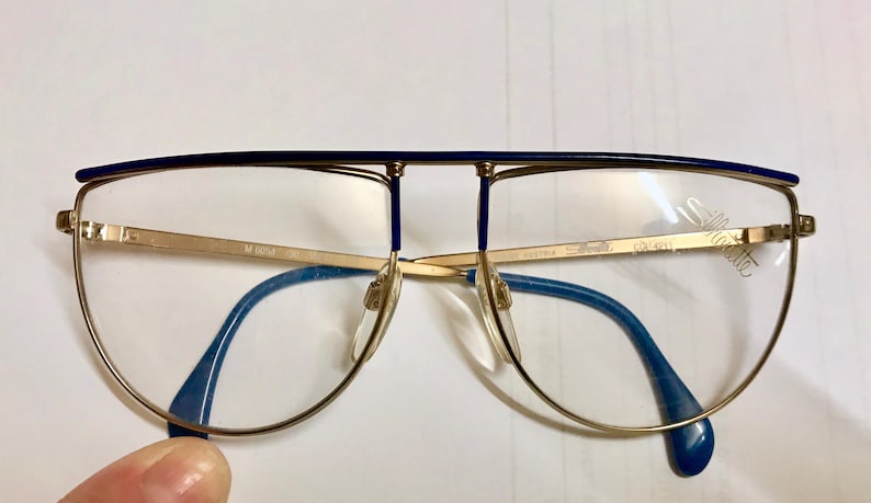 May include: Gold-rimmed eyeglasses with a blue top bar and blue temples. The lenses are clear, and the frame has a distinctive aviator shape. The brand name "Silhouette" is visible on the lens.