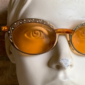 Op de afbeelding: Ovale zonnebril met amberkleurige glazen en goudkleurige metalen monturen. De bovenrand van de monturen is versierd met een herhalend geometrisch patroon van ge&euml;tste lijnen en kleine, heldere kristallen die langs de bovenkant zijn ingelegd.