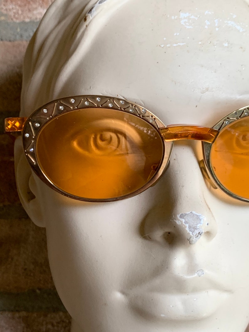Op de afbeelding: Ovale zonnebril met amberkleurige glazen en goudkleurige metalen monturen. De bovenrand van de monturen is versierd met een herhalend geometrisch patroon van ge&euml;tste lijnen en kleine, heldere kristallen die langs de bovenkant zijn ingelegd.