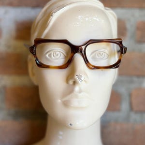 Puede incluir: Un par de gafas rectangulares con una montura de patrón de carey marrón oscuro. Las gafas presentan un puente angular distintivo y bordes gruesos, colocadas sobre una cabeza de maniquí lisa y clara con rasgos faciales definidos.