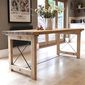 Op de afbeelding: Een lichtgekleurde houten eettafel met een rechthoekig blad en vier stevige poten. De tafel heeft zwarte metalen kruissteunen voor extra stabiliteit. Een vaas met witte bloemen en kleine bruine vazen staan op de tafel. De tafel is ongeveer 183 cm lang.