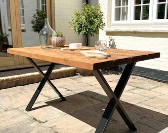 May include: A wooden dining table with a natural finish and black metal X-shaped legs. The table is set with a glass vase, a small potted plant, a white plate, and a wooden chopping board.