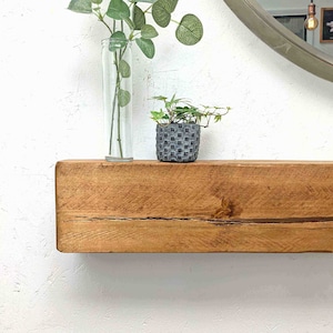 Op de afbeelding: Een rustiek houten plank met een natuurlijke houtkleur. De plank is gemonteerd aan een witte muur en heeft twee planten in potten erop. De planten staan in een glazen vaas en een grijze keramische pot.
