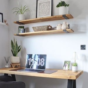 May include: Two wooden shelves with black metal brackets mounted on a white wall. The shelves are decorated with plants, a framed print, and decorative pottery. A desk with a laptop, a lamp, and a picture frame sits below the shelves.