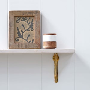 May include: A framed print with a floral design, a small wooden container with a white band, and a brass shelf bracket. The items are displayed on a white shelf against a white wall. The frame and container are made of wood.