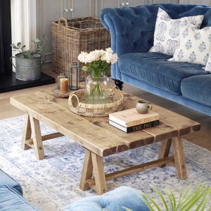 Könnte beinhalten: Ein rechteckiger Couchtisch aus hellem Holz mit einem rustikalen Design und einem Bockfuß. Auf dem Tisch befinden sich ein geflochtener Korb, Bücher und eine Tasse. Im Hintergrund ein blaues Samtsofa und gemusterte Kissen.