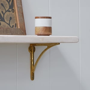 May include: A shelf supported by a gold-coloured metal bracket. A wooden and white ceramic container sits on the shelf. A framed picture is visible on the left. The shelf is mounted on a white wall.